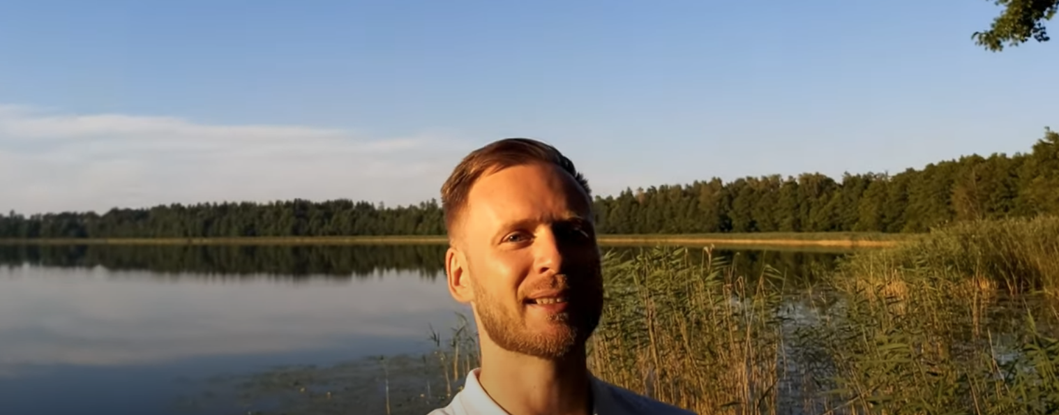 Mężczyzna w białej koszulce stoi nad jeziorem w otoczeniu zieleni, w tle widoczne drzewa i błękitne niebo.