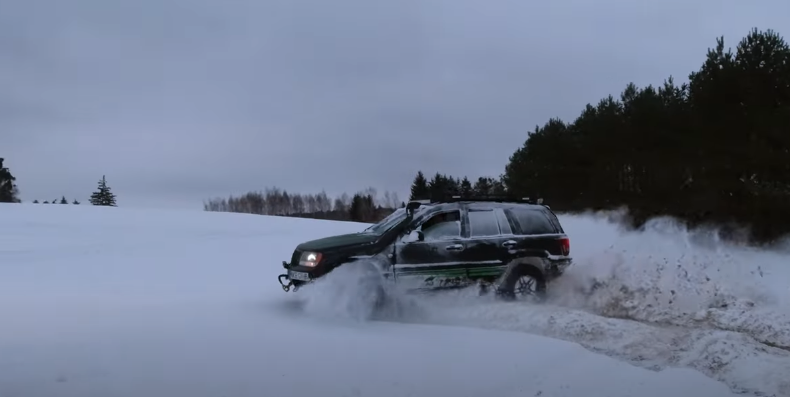 Samochód terenowy ściga się po zaśnieżonym terenie, tworząc chmurę śniegu. W tle widoczne drzewa, a niebo jest pochmurne. Idealna scena do off-roadu w zimowych warunkach.