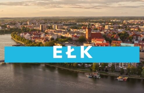 Widok na miasto Ełk z lotu ptaka, z rzeką Ełk w pierwszym planie, otoczone malowniczymi budynkami i zielenią.