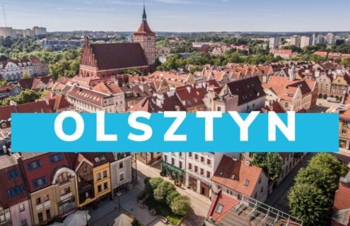 Widok panoramiczny na Olsztyn, z charakterystycznymi czerwonymi dachami budynków oraz zielonymi terenami. W tle widać gotycki zamek oraz nowoczesne budynki miasta.