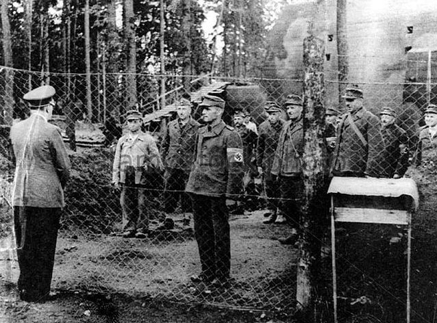 Czarno-biała fotografia przedstawiająca żołnierzy stojących za siatką w otoczeniu lasu.