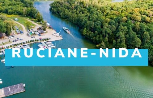 Widok lotniczy na Ruciane-Nidę, malowniczą miejscowość w Polsce, otoczoną jeziorami i lasami. Widać port, przystań oraz łodzie na wodzie, co sugeruje możliwość uprawiania sportów wodnych i rekreacji na świeżym powietrzu.
