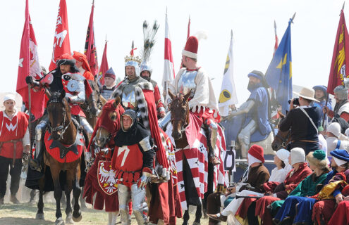 Uczestnicy rekonstrukcji historycznej w średniowiecznych strojach, jeżdżący na koniach, otoczeni flagami i sztandarami. Na pierwszym planie rycerze w zbrojach, w tle grupy ludzi w tradycyjnych strojach.